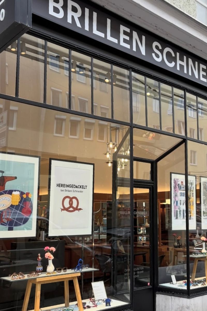 Schaufenster bei Brillen Schneider, Optiker in München, mit vonSUSI Ausstellung – farbenfrohe Poster mit Humor, Typografie und bayerischem Lebensgefühl.